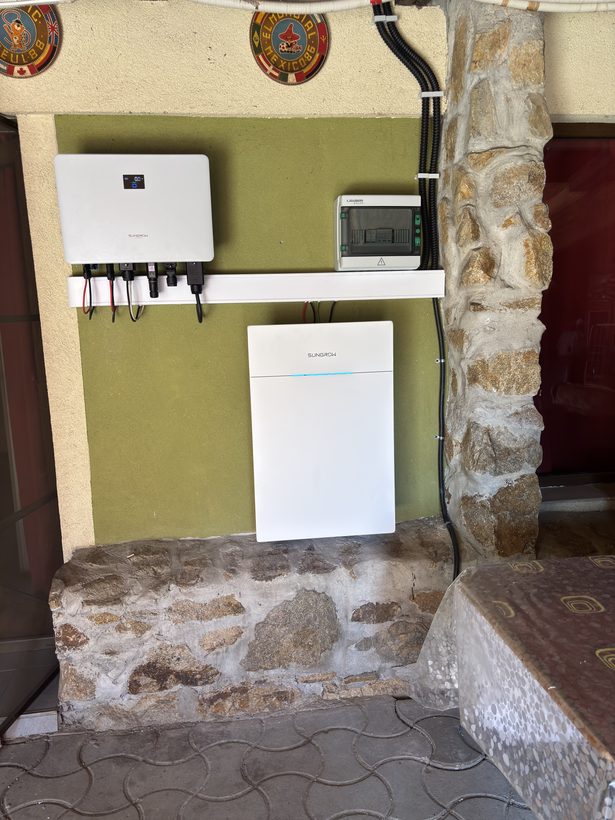 Invertor Sungrow, tablou de protecție și baterie Sungrow montate pe un perete verde de terasă, lângă un stâlp din piatră